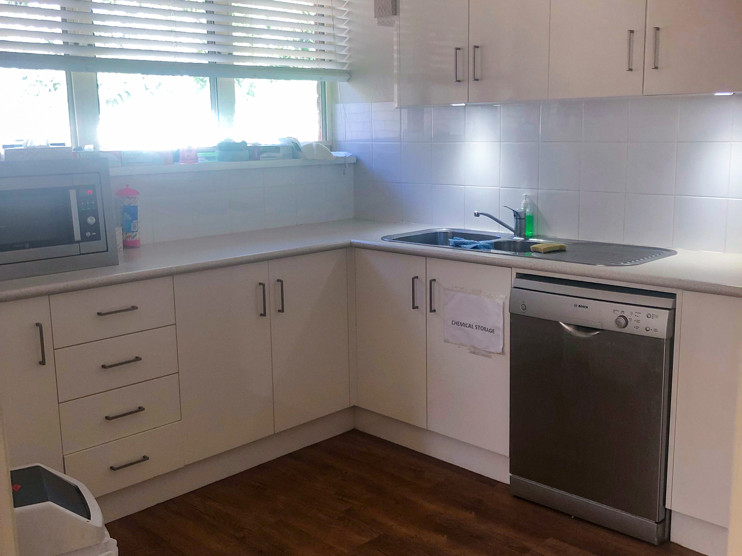 Kitchen with off-white cabinetry, white tiled splash backs and timber look flooring, A large dishwasher, sink and microwave in stainless steel are visibile