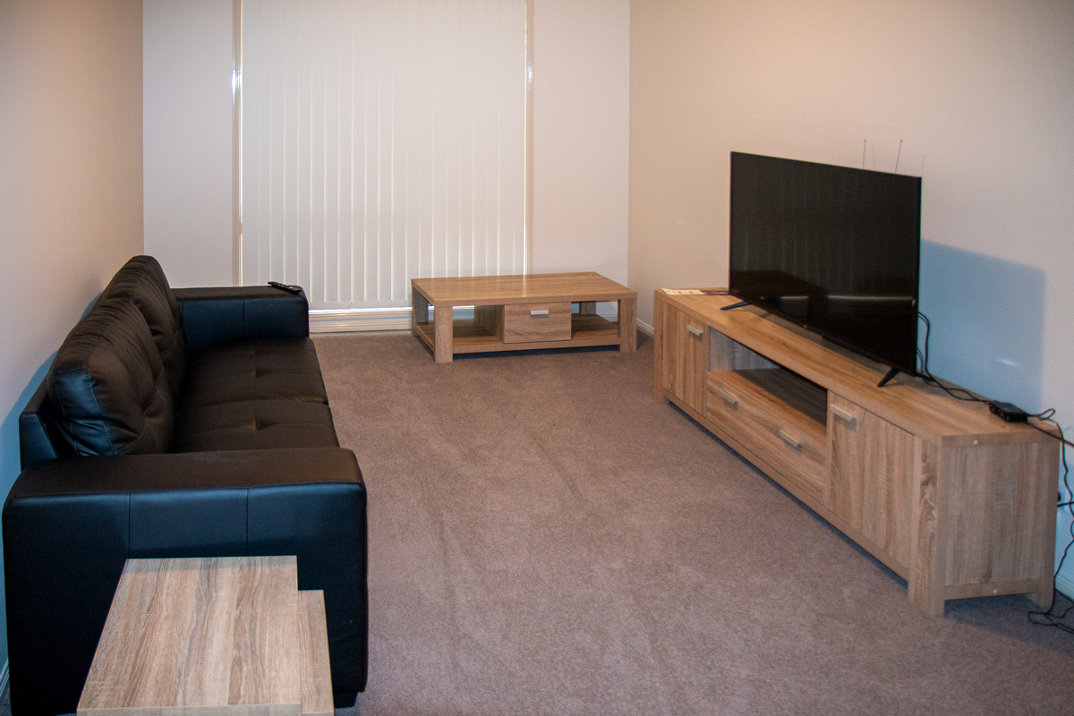A cozy carpeted lounge room with timber entertainment unit and matching coffee table and side table. A large black leather lounge sits opposite the TV