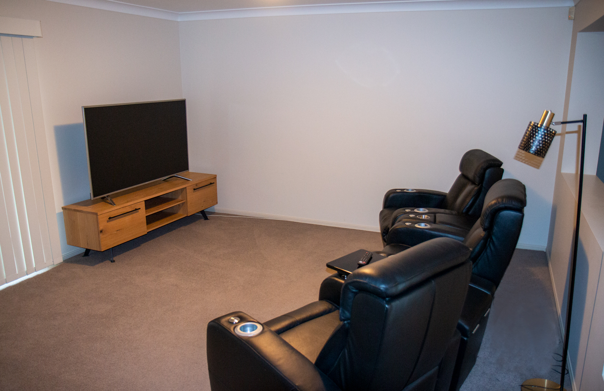 Carpeted living room with entertainment unit with a TV sitting on top. 3 black leather reclining lounge chairs face the TV.