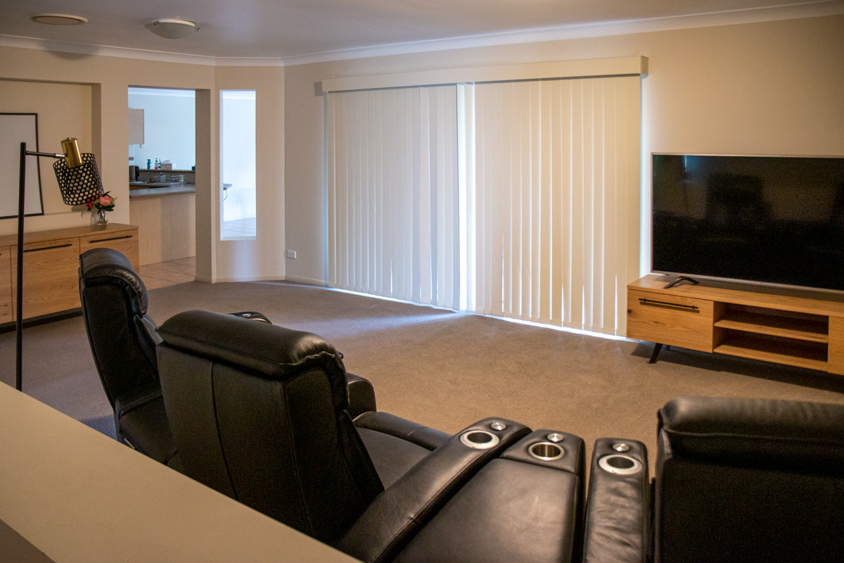 View from the living room into the kitchen. 3 leather reclining lounge chairs and the TV and entertainment unit are in the foreground. 2 large glass doors with vertical blinds are on the far wall which wraps around to the kitchen area