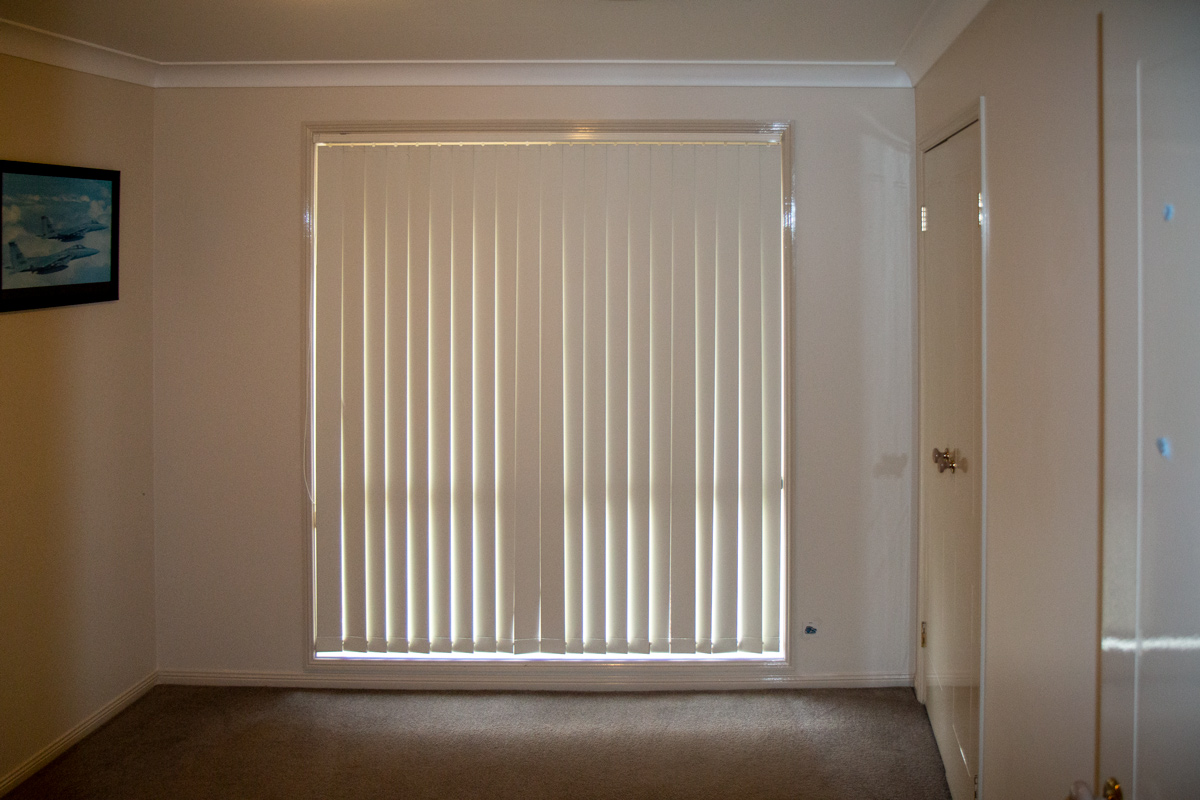 A vacant bedroom with carpeted floors and a large window with vertical blinds. A 2 door built in wardrobe can be seen in the right hand corner