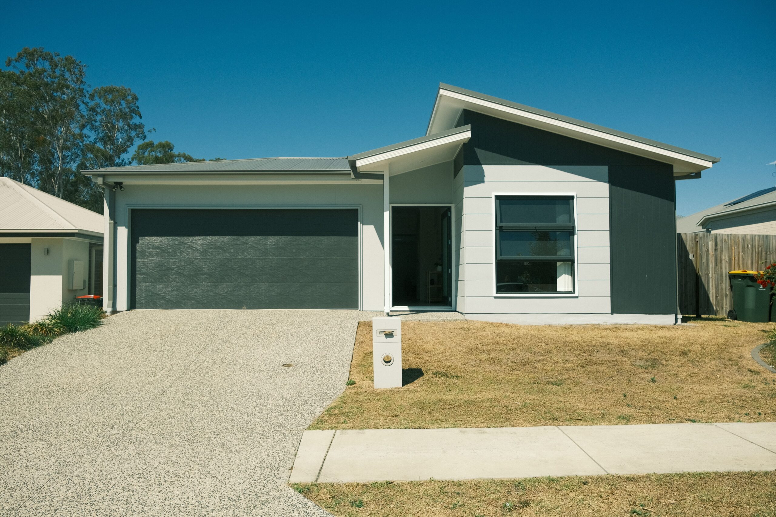 Front of Home - there is a driveway sloping upwards towards a garage on the left of the image. On the right, there is the front door and a bedroom window. There is a small front yard with a lawn.