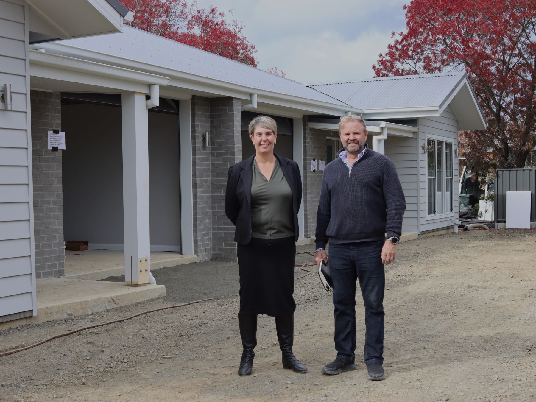A woman and a aman stand outside newly built SDA homes. The homes are grey and there are brightly coloured red autumn trees in the background behind them.