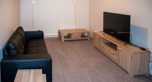 A cozy carpeted lounge room with timber entertainment unit and matching coffee table and side table. A large black leather lounge sits opposite the TV