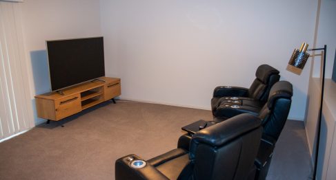 Carpeted living room with entertainment unit with a TV sitting on top. 3 black leather reclining lounge chairs face the TV.