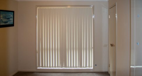 A vacant bedroom with carpeted floors and a large window with vertical blinds. A 2 door built in wardrobe can be seen in the right hand corner