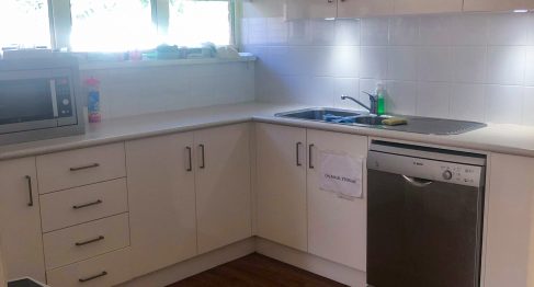 Kitchen with off-white cabinetry, white tiled splash backs and timber look flooring, A large dishwasher, sink and microwave in stainless steel are visibile