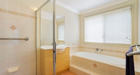 Interior bathroom. Built in tiled bath below a large window with blinds, tiled floors, a small vanity and wash basin with a large mirror and a large easy access shower stall with grab rails.
